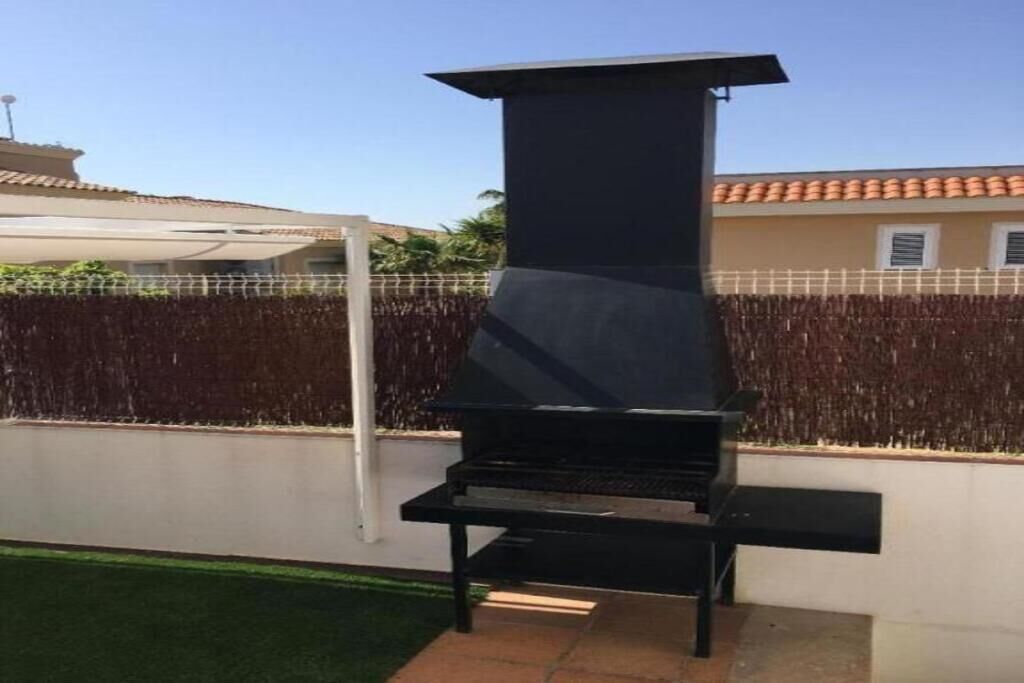 A black outdoor grill with a chimney, set against a white wall and a fence.