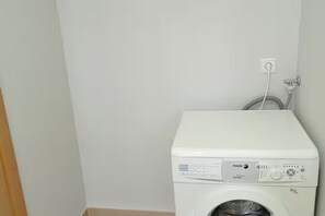 Eine weiße Waschmaschine steht in einer Ecke des Raumes, an einer weißen Wand mit einer sichtbaren Steckdose.