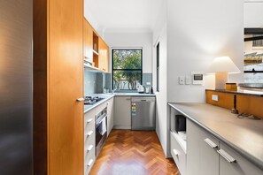 A narrow kitchen with wooden cabinets, a herringbone floor, and a window with a view of trees. - Riley Escape Walkable City Living with Courtyard (Surry Hills)