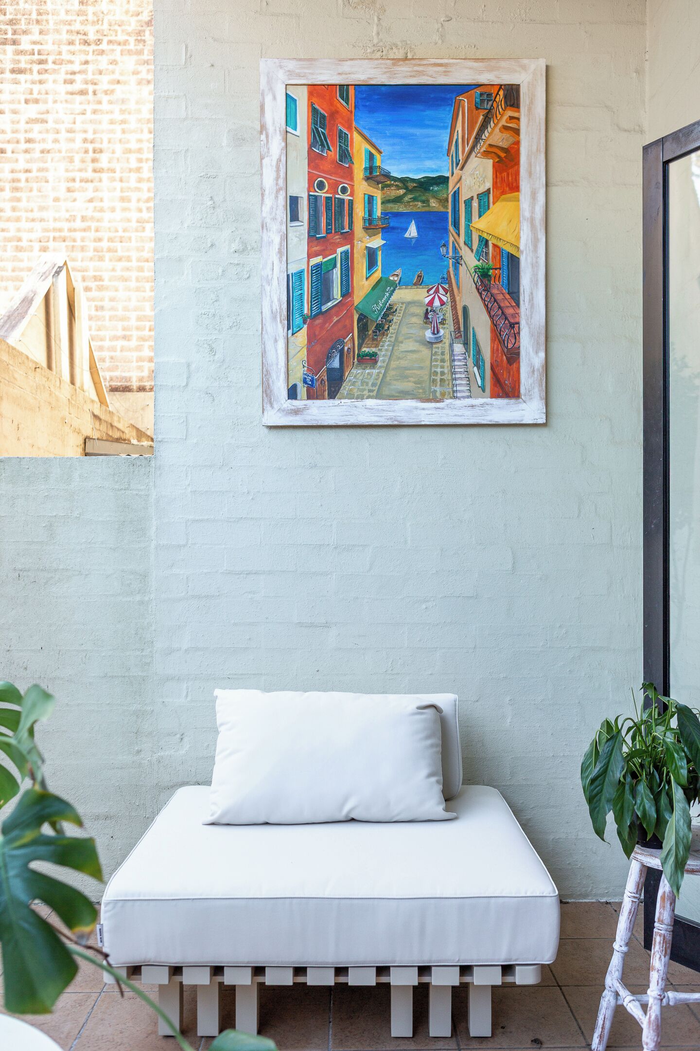 A room with a bed, a painting of a coastal street, and a plant.