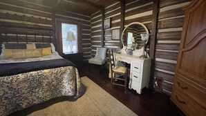 A log cabin bedroom with a bed, dresser, chair, and a window with a view.