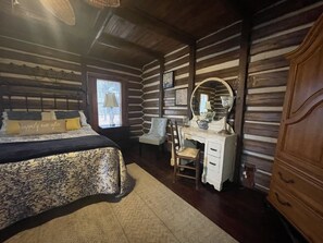 A log cabin bedroom with a bed, dresser, chair, and a window with a view.