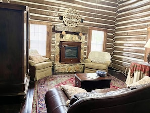 A log cabin interior with a fireplace, leather sofa, armchairs, and a patterned rug.