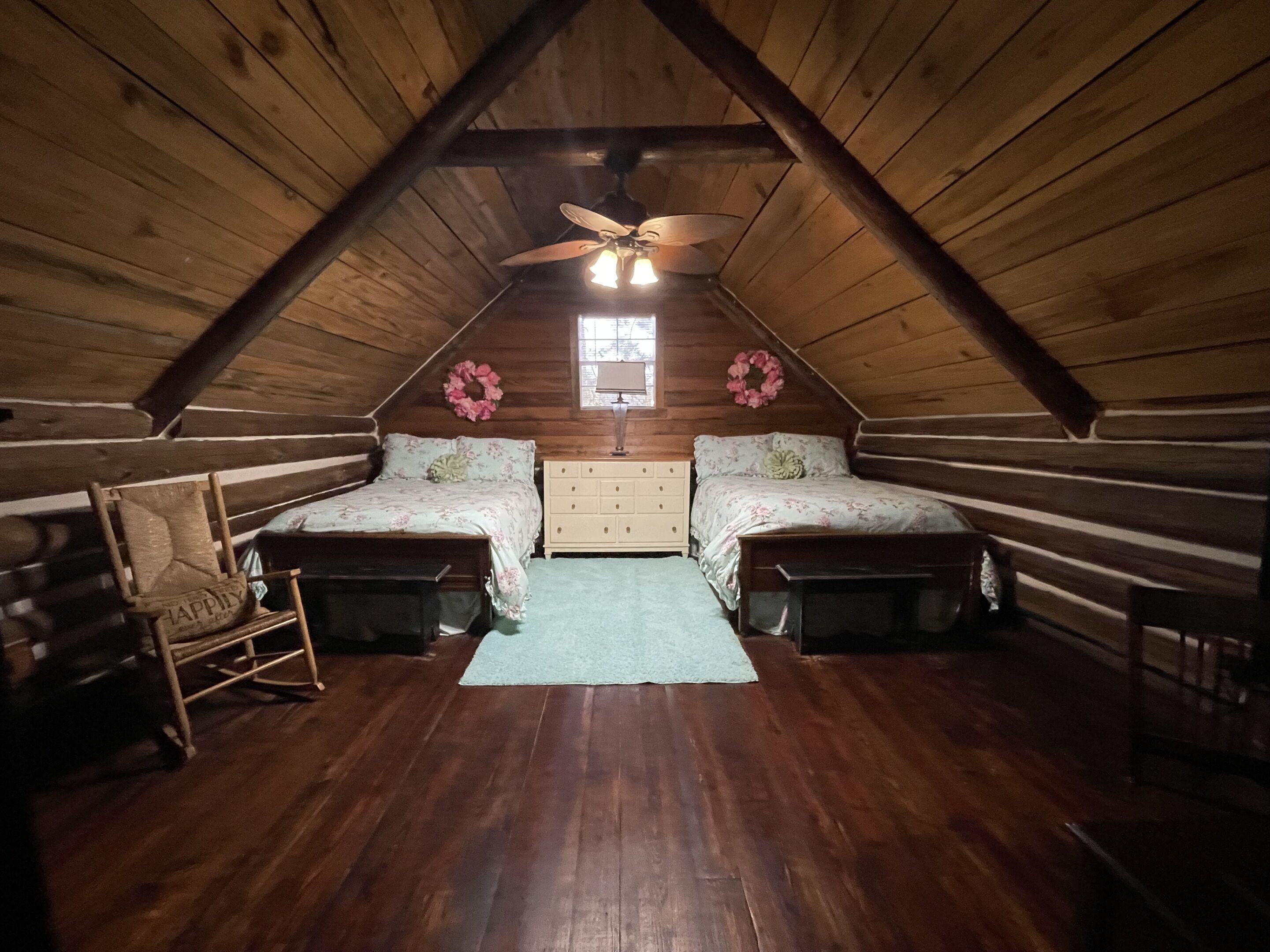 A log cabin bedroom with two beds, a dresser, a rocking chair, and a ceiling fan.