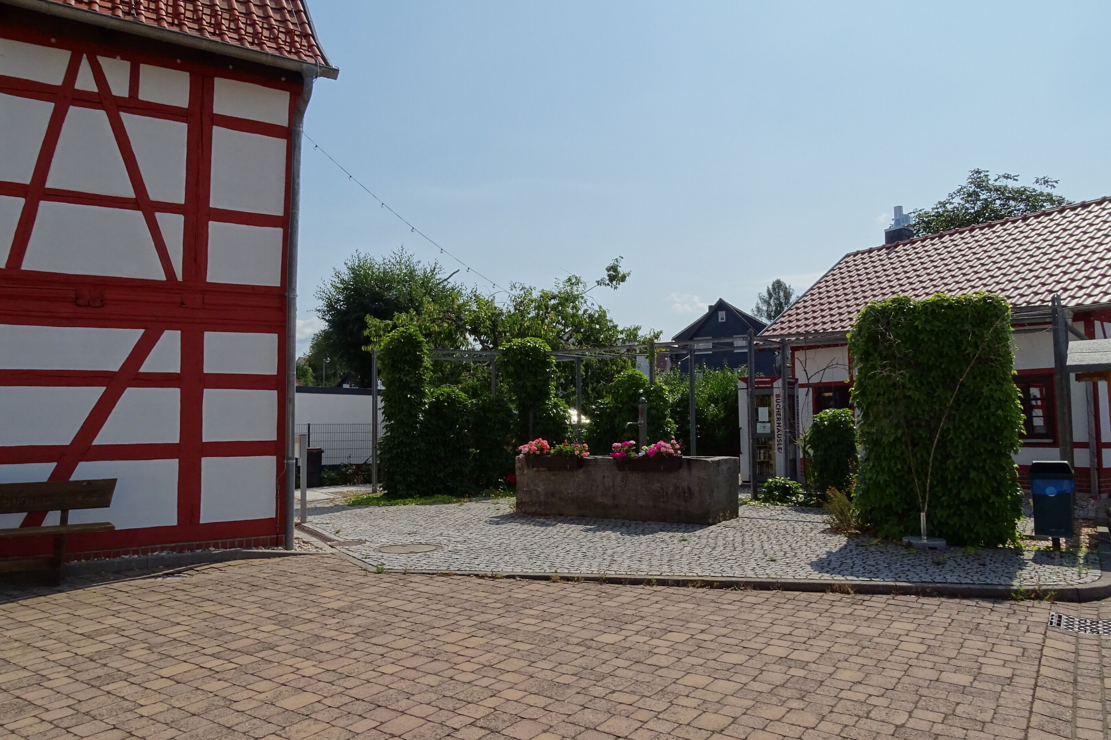 Une cour dallée de pierres, un bâtiment traditionnel à colombages et un parterre de fleurs roses.