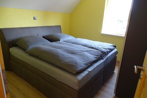 Ein Einzelbett mit grauem Kopfteil und zwei Kissen, ein Holzboden und ein Fenster mit Tageslicht.