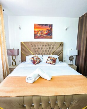 Une chambre d’hôtel avec un grand lit, une tête de lit à motifs capitonnés, deux coussins décoratifs, un tableau représentant des lions et une serviette pliée posée sur le lit.