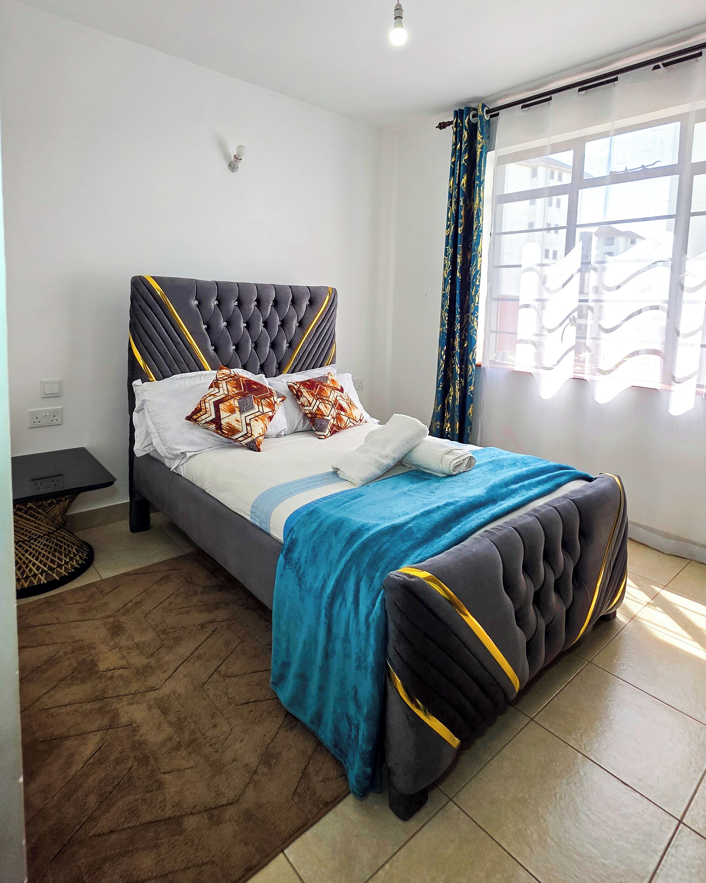 Un dormitorio moderno con un cabecero capitonado, una cama con almohadas estampadas, una manta azul y vistas al exterior a través de una ventana con cortinas estampadas.