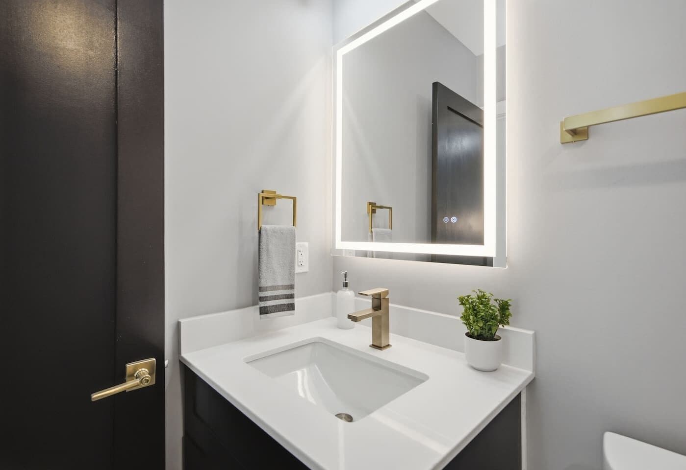 Ein modernes Badezimmer mit einem weißen Waschbecken, einem goldenen Wasserhahn, einem Handtuch an einem goldenen Halter und einem Spiegel mit Beleuchtung darüber.