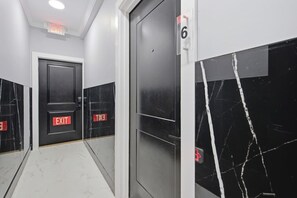 Um corredor com paredes de mármore preto, duas portas com placas de saída vermelhas e teto branco com sinalização de emergência.