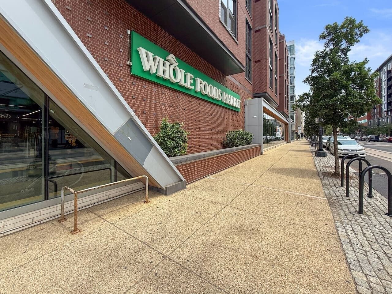 Un magasin Whole Foods Market avec une façade en briques, une entrée bien visible et un trottoir bordé d’une barrière métallique.