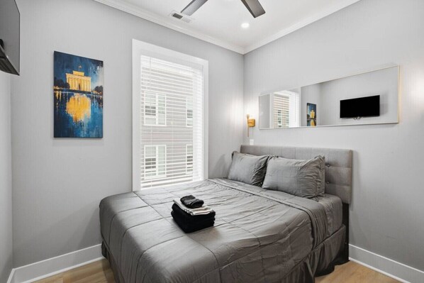 A bedroom with a bed, a window with blinds, a painting, and a television on the wall.