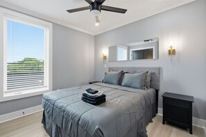 Ein Schlafzimmer mit einem Bett, Deckenventilator, Nachttisch, Kommode und einem Fenster mit Jalousien.