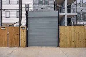 Una entrada protegida por un portón enrollable gris, flanqueada por cercas de madera, que conduce a un edificio con múltiples ventanas y balcones.