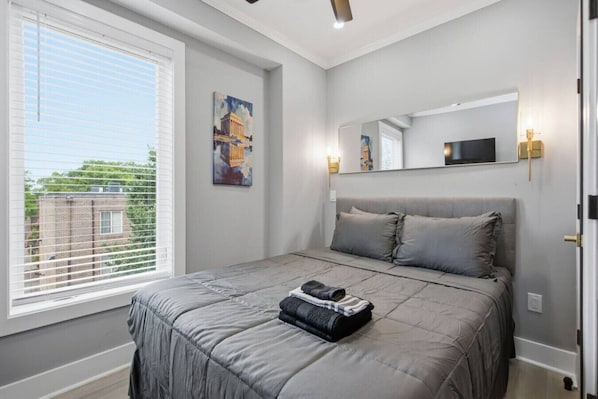 Une chambre à coucher avec un lit, une fenêtre avec des stores, un tableau et un miroir.