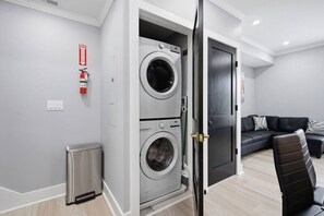 Ein moderner Waschraum mit zwei übereinander gestapelten Waschmaschinen, einem Feuerlöscher, einer kleinen Mülltonne und einem dunkelgrauen Sofa in einem Wohnbereich.
