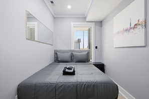 Een slaapkamer met een bed, nachtkastje, spiegel en raam met jaloezieën.