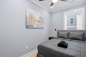 Een slaapkamer met een bed, een raam met jaloezieën, een plafondventilator en een schilderij aan de muur.