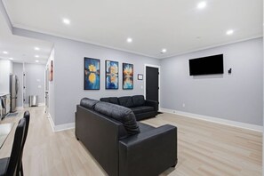 A modern living room with a sectional sofa, a flat-screen TV mounted on the wall, and a dining area with a table and chairs.