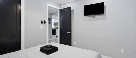 Un dormitorio con una cama, ventilador de techo, televisor montado en la pared y una puerta que da a una cocina.