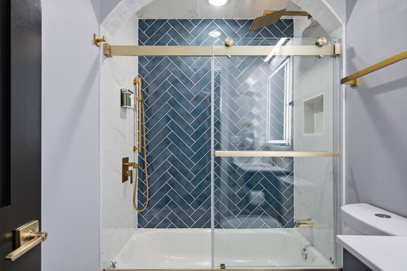 A modern bathroom with a glass-enclosed shower, a white bathtub, and a herringbone-patterned tile wall.
