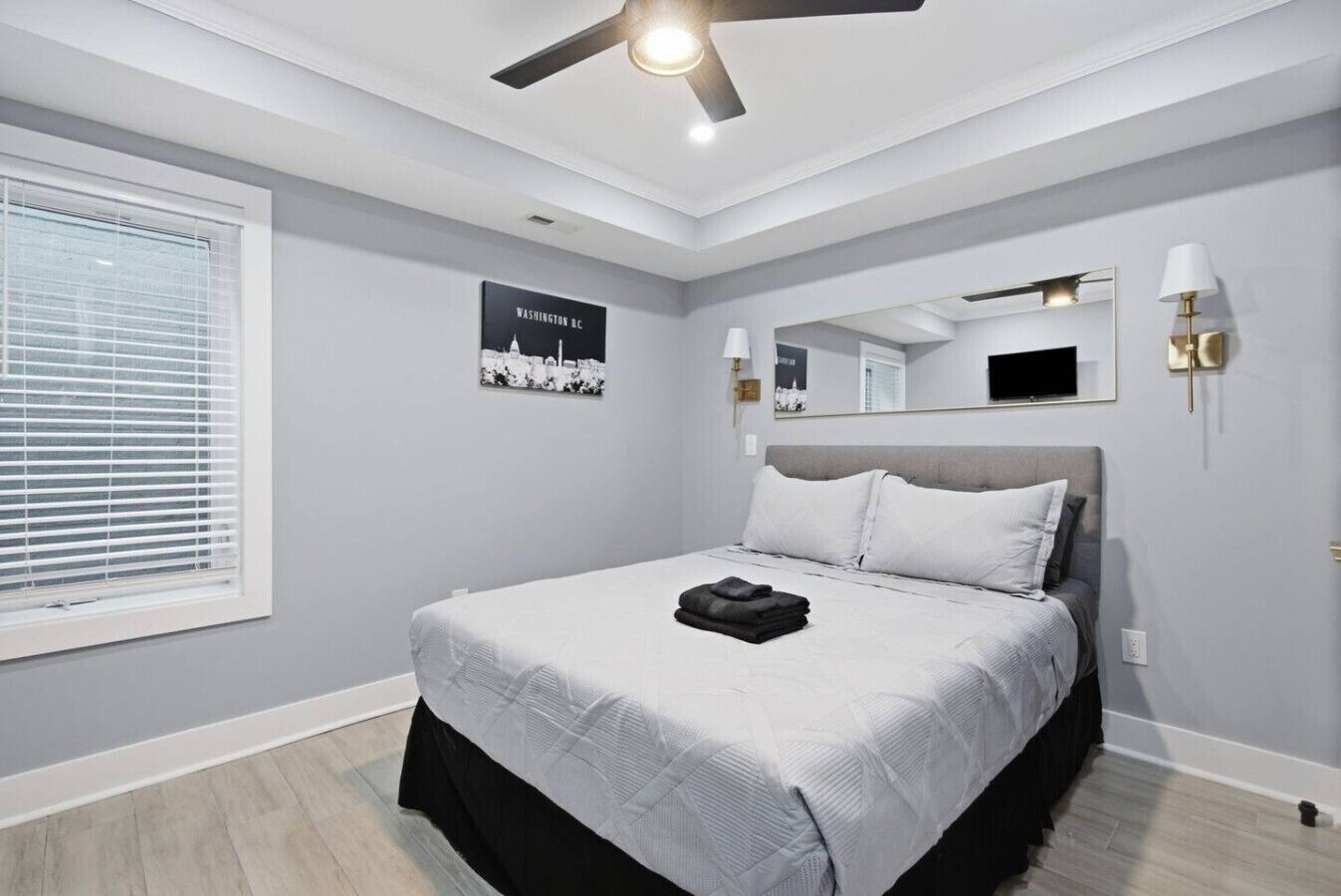 A bedroom with a bed, a ceiling fan, a window with blinds, a wall-mounted TV, and a poster.