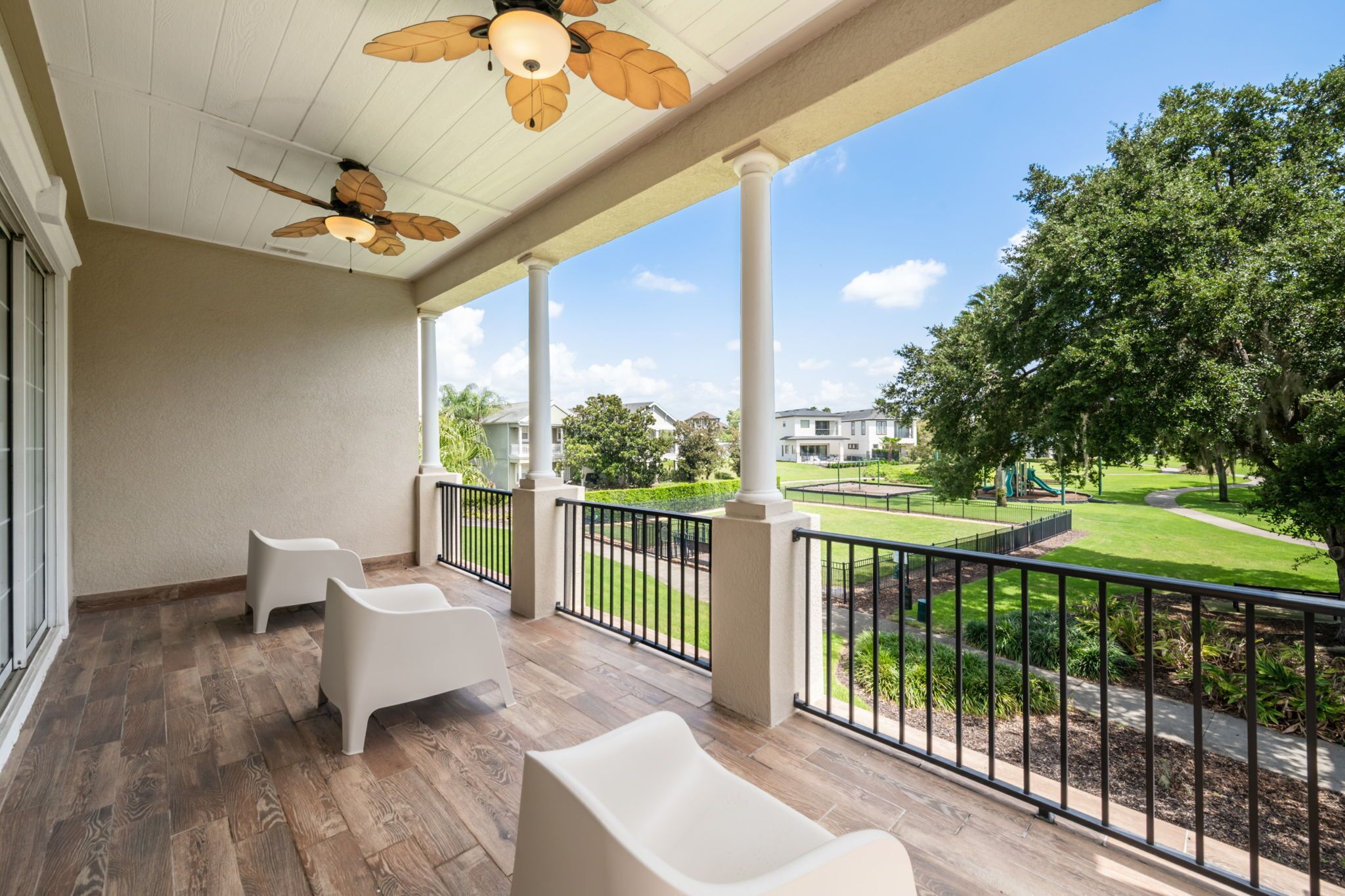 A covered outdoor area with a ceiling fan, white chairs, and a view of a residential neighborhood.