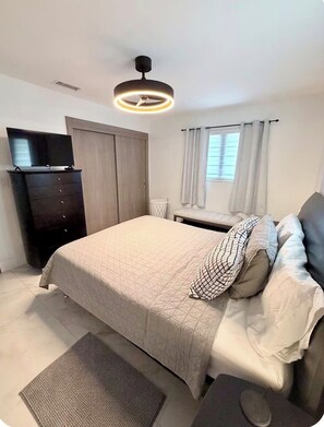 Une chambre à coucher avec un lit, un téléviseur posé sur une commode, un ventilateur de plafond et une fenêtre avec des rideaux.