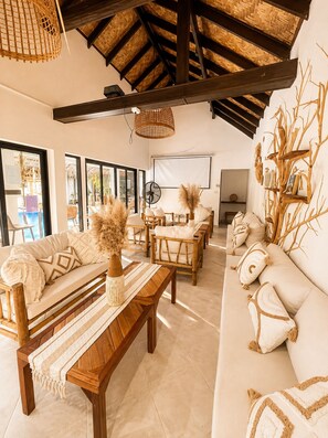 A room with a high ceiling, wooden beams, a sofa set, a bench, and a wall-mounted shelf with decorative items. - Bohemian-style 4-bedroom villa in Bali with private pool, sauna, and ice bath; beach just a 1-minute walk away (Surat Thani)