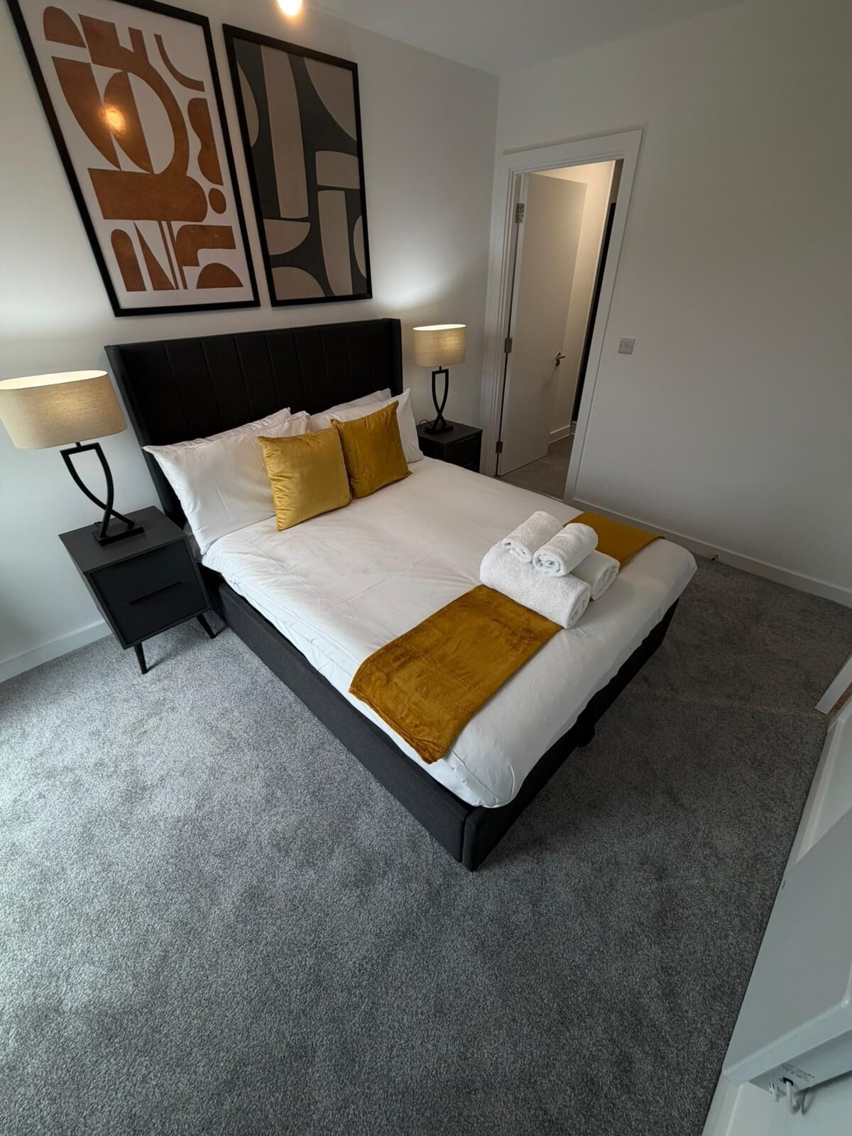 Un lit bien fait, avec du linge de lit blanc, deux coussins dorés et un plaid marron. On y trouve deux tables de chevet avec des lampes, une porte donnant sur une autre pièce et, au-dessus du lit, des œuvres d’art abstraites accrochées au mur.