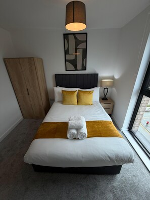 Ein ordentlich bezogenes Bett mit weißer und goldener Bettwäsche, ein Holzschrank, ein moderner Kopfteil, ein Wandbild und ein Fenster mit Blick auf die Straße.