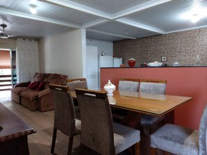 A dining area with a wooden table and chairs, a sofa with cushions, and a kitchen area with a refrigerator and decorative items.