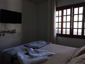A room with a bed, a flat-screen TV mounted on the wall, and a window with a view of the outside.