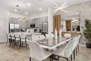 A modern kitchen with a central island, white cabinetry, stainless steel appliances, and a dining area with a long table and chairs. - The Premiere House - 8 Mins to Universal Studios 3 Mins to Sea World 15 Mins to Disney World (Orlando)