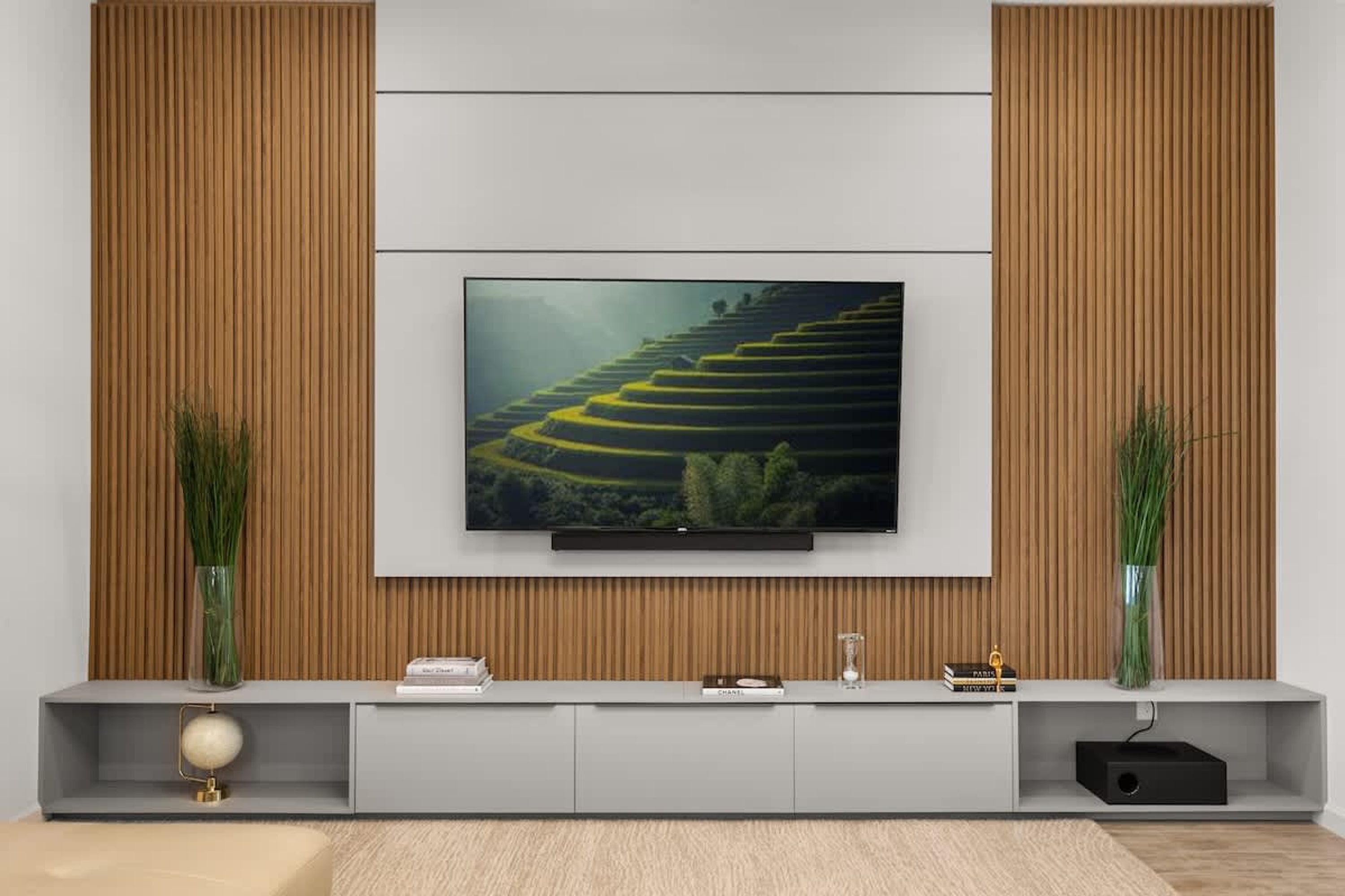 Una sala de estar moderna con un televisor de pantalla plana montado en una pared de madera, un mueble de entretenimiento gris debajo y dos jarrones altos con plantas verdes.