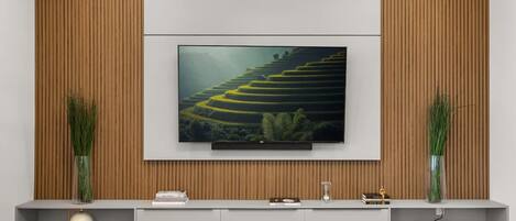 A modern living room with a flat-screen TV mounted on a wooden wall, a grey entertainment unit below, and two tall vases with green plants.