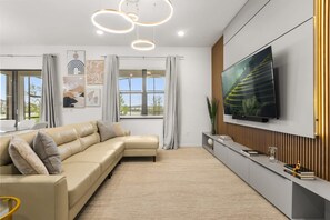 A modern living room with a sectional sofa, a flat-screen TV mounted on the wall, and a large window with curtains. - The Premiere House - 8 Mins to Universal Studios 3 Mins to Sea World 15 Mins to Disney World (Orlando)