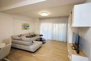 A modern living room with a sofa, a coffee table, a painting on the wall, a television, and a window with curtains. - Luxury Athens Duplex with Acropolis View, Private Heated Jacuzzi & Parking (Athina)