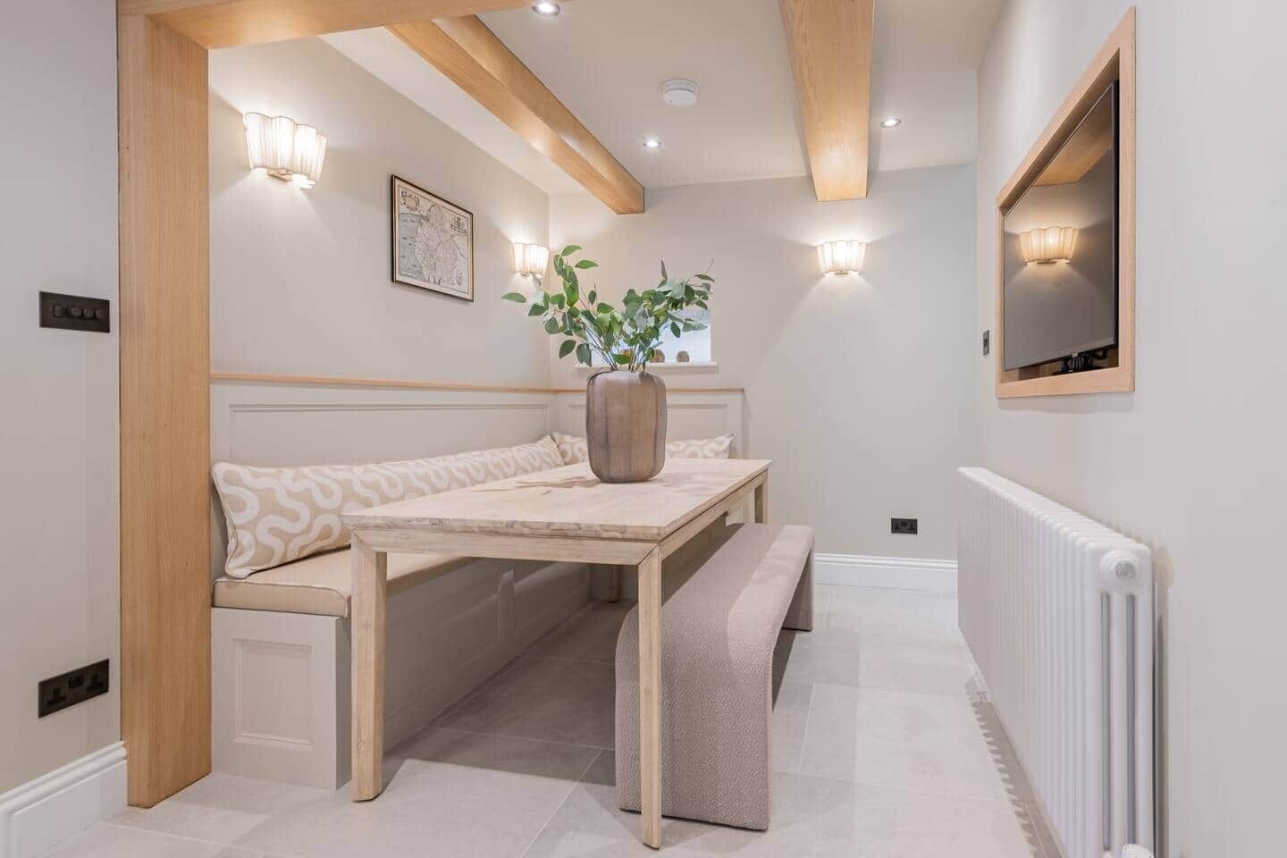 A room with a bench, a table, a potted plant, a wall-mounted TV, and recessed lighting.