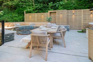 An outdoor patio with a stone fire pit, wooden furniture, and a dining table set for four. - Easter Crag, 3 bedroomed luxury escape with hot tub (Bowness-on-Windermere)