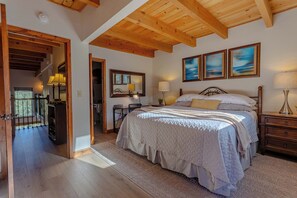 A bedroom with a large bed, wooden ceiling beams, a desk with a chair, and a view of the outdoors.