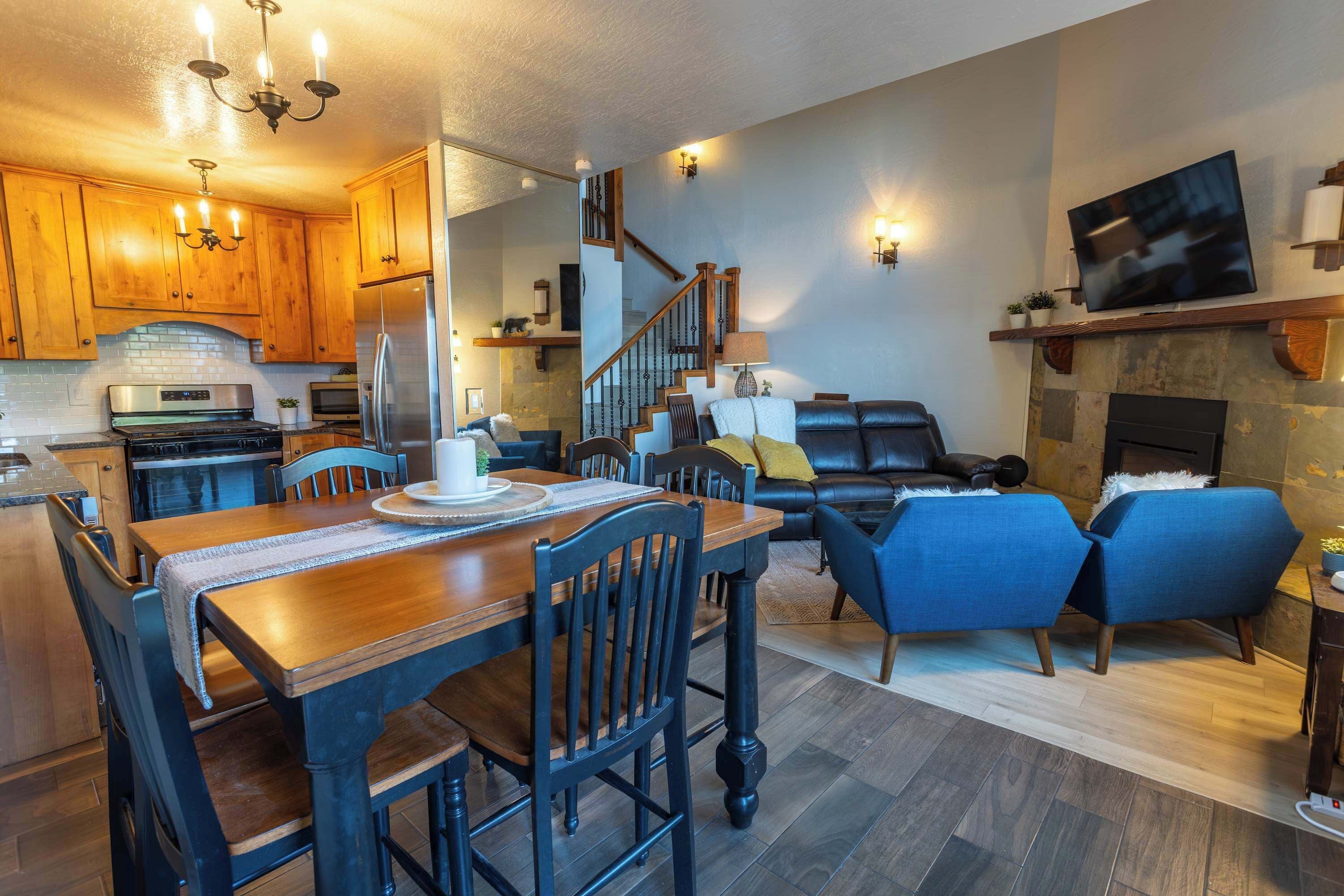 A modern kitchen with wooden cabinets, a dining table with chairs, a living area with a sofa and chairs, and a fireplace.