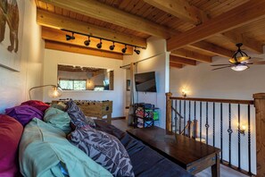 Una sala de estar acogedora con techo de madera, un sofá con cojines estampados, una mesa de centro de madera y una escalera con barandilla.