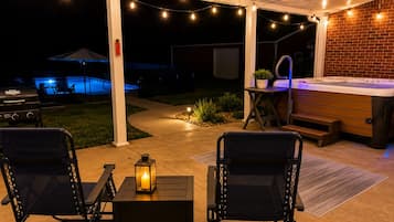 Uma área de estar ao ar livre com hidromassagem, duas cadeiras, uma mesa com um candeeiro e luzes de corda.