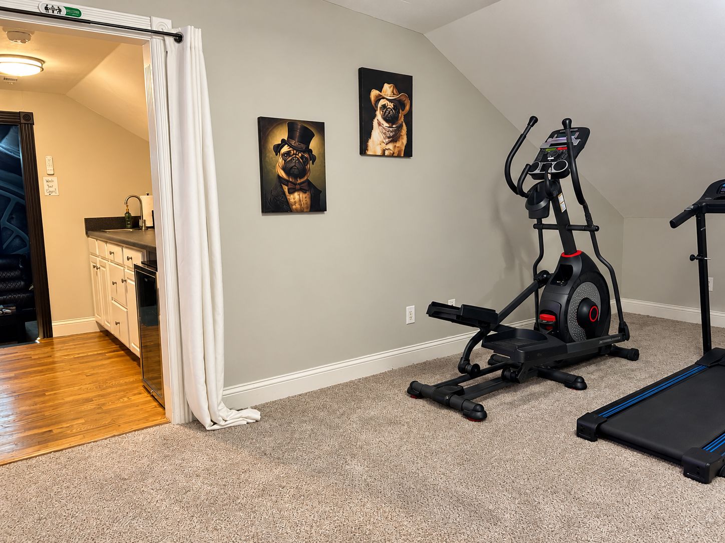 Un gimnasio en casa con equipo de ejercicio, una zona de cocina y dos cuadros en la pared.