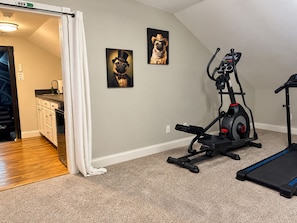 A home gym with exercise equipment, a kitchen area, and two framed pictures on the wall.