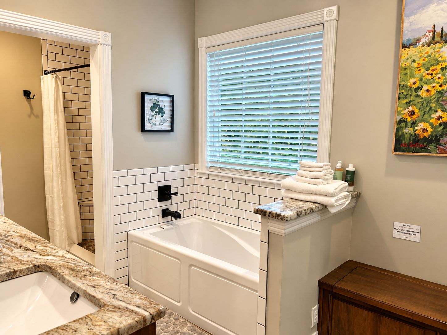 A bathroom with a bathtub, a sink, a window with blinds, a towel rack, and a painting on the wall.