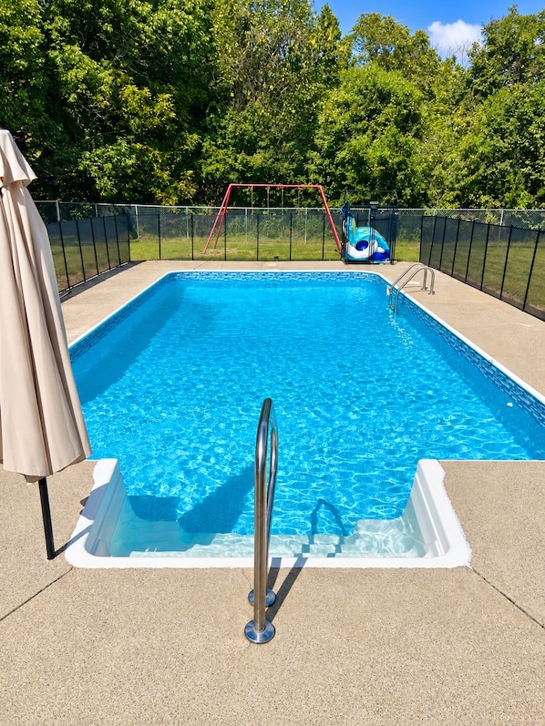 A rectangular swimming pool with a metal ladder, surrounded by a fence, and a slide in the background.