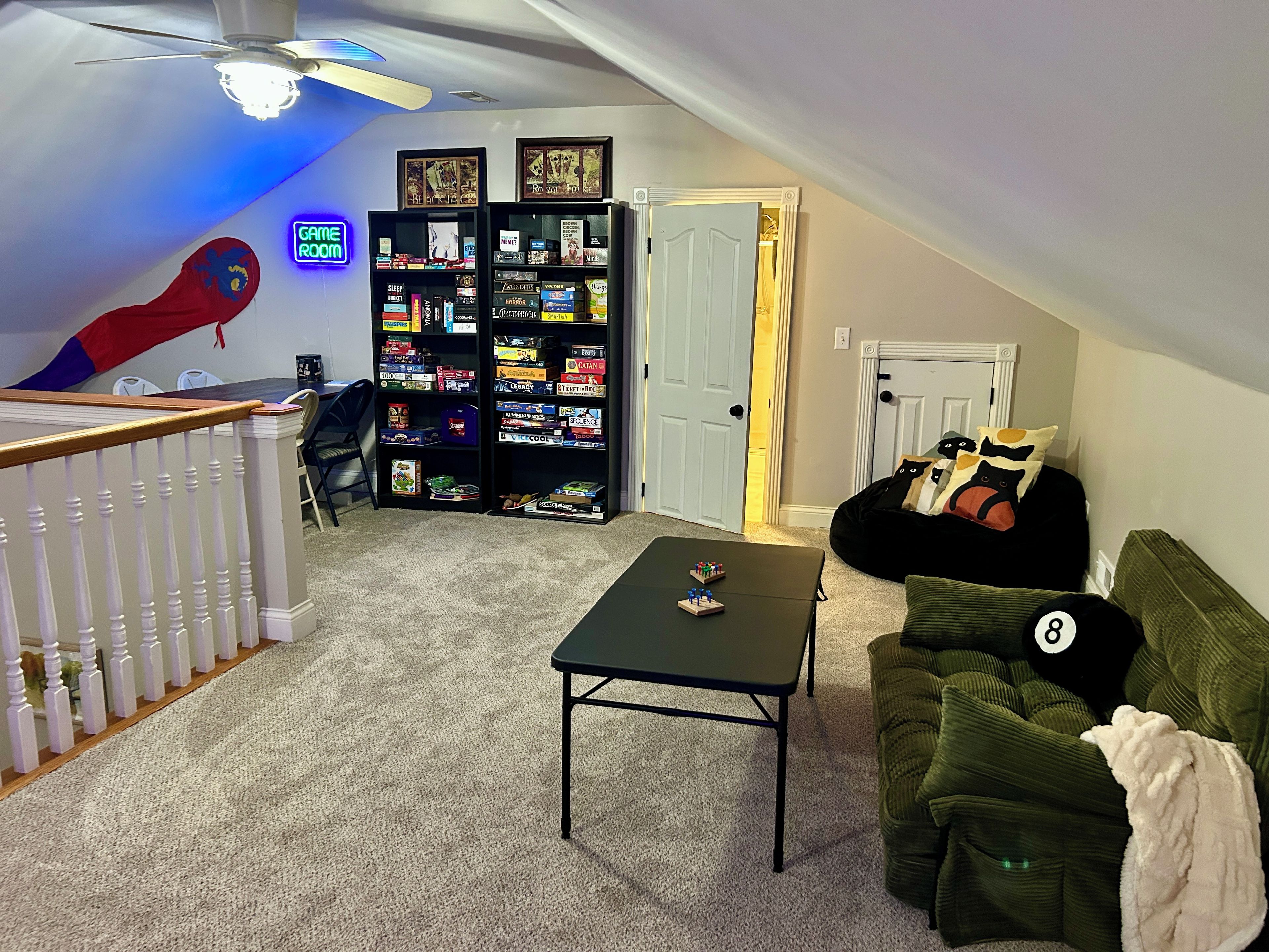 Um quarto no sótão com teto inclinado, ventilador de teto, estante para jogos, sofá verde e uma pequena mesa.