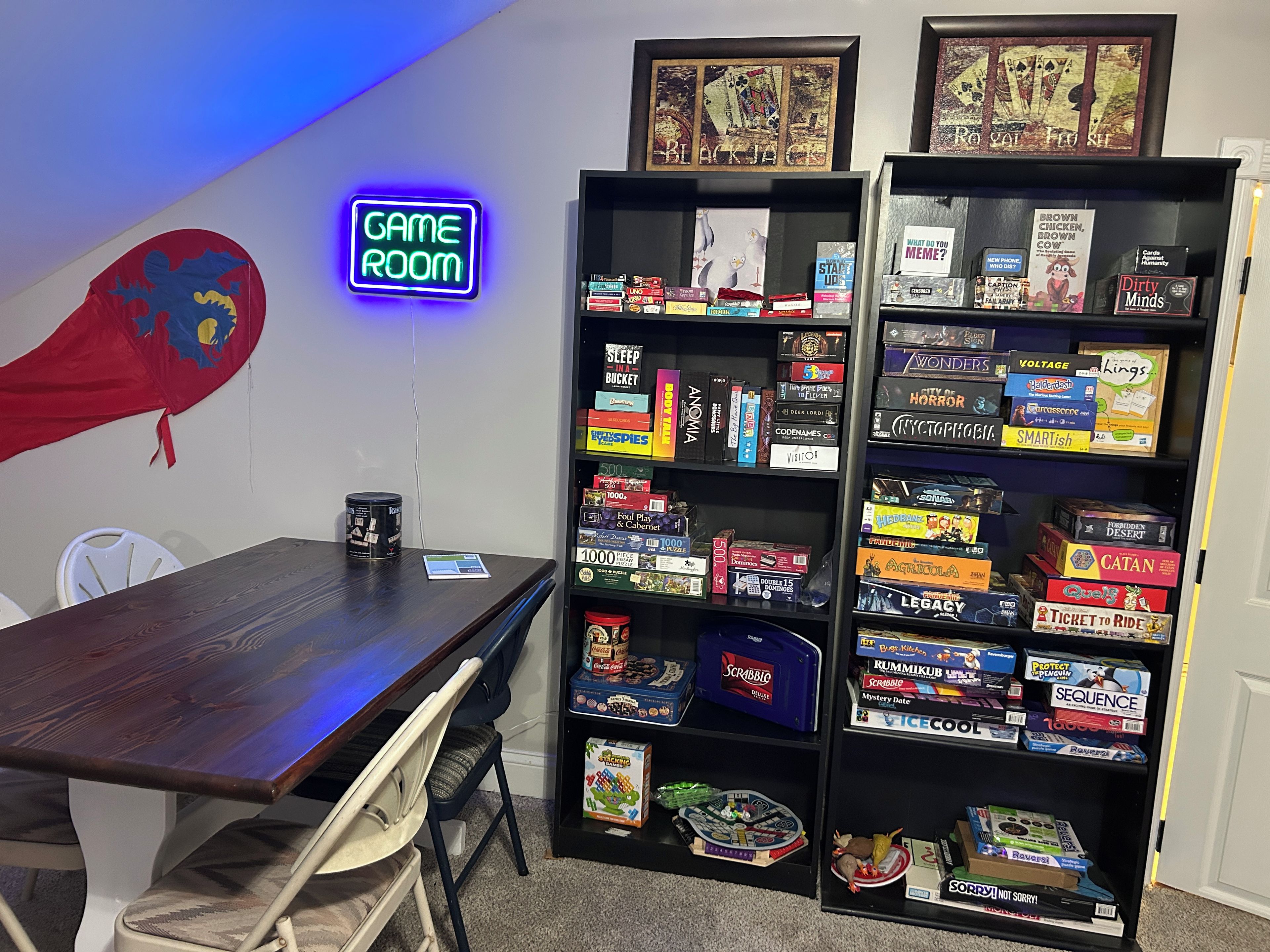 Una habitación con una mesa de madera, un letrero de neón que dice 'GAME ROOM' y estantes llenos de juegos de mesa.